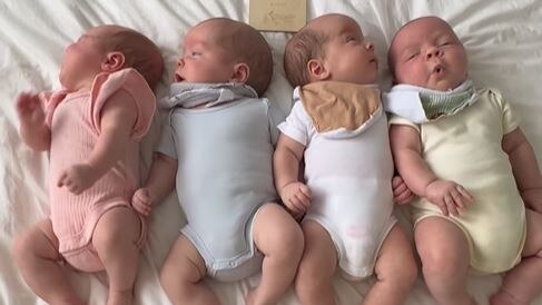 4 white babies on a white bed sheet. They are each wearing differently coloured pastel onesies.