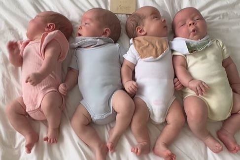 4 white babies on a white bed sheet. They are each wearing differently coloured pastel onesies.