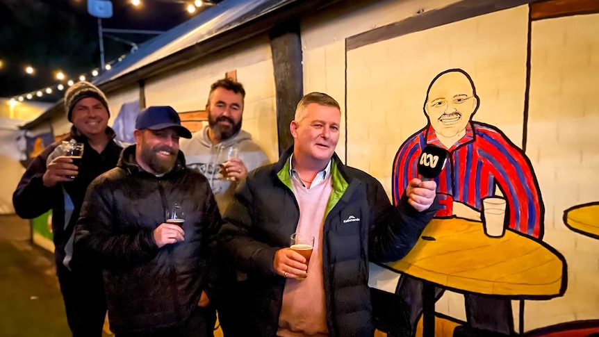 Four men stand next to a colourful mural of a smiling man seated drinking a pint.