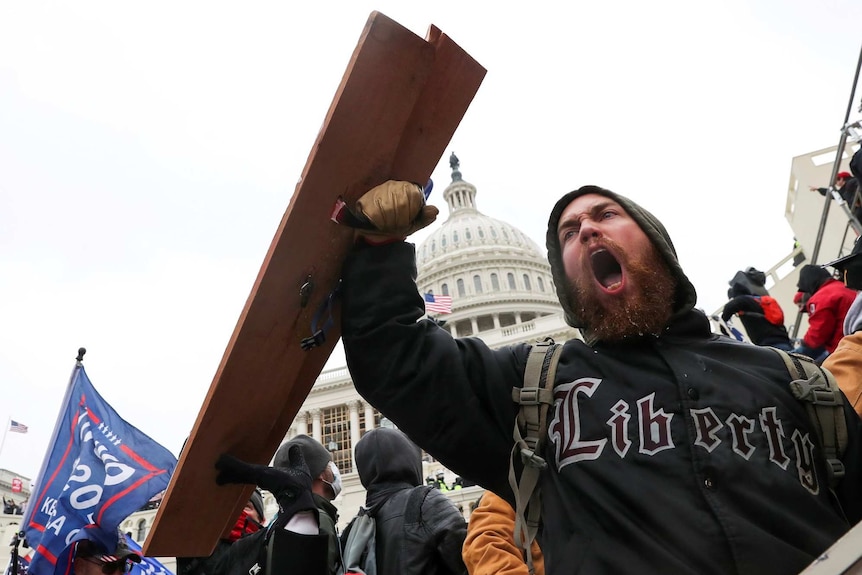 A man in a shirt that says 'liberty' holds his fist in the air and shouts with the Capitol building behind him