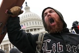 A man in a shirt that says 'liberty' holds his fist in the air and shouts with the Capitol building behind him