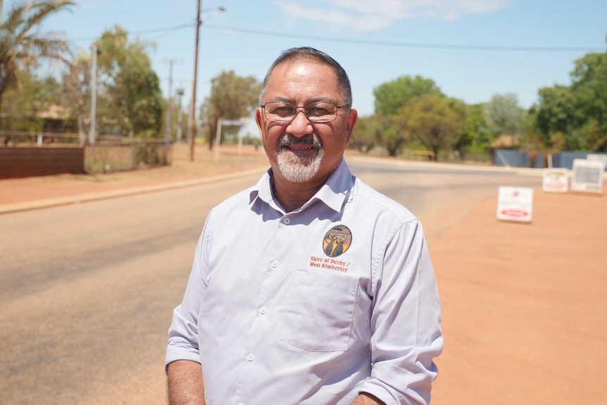 An older man with a white beard stands smiling on a road in a desert town.