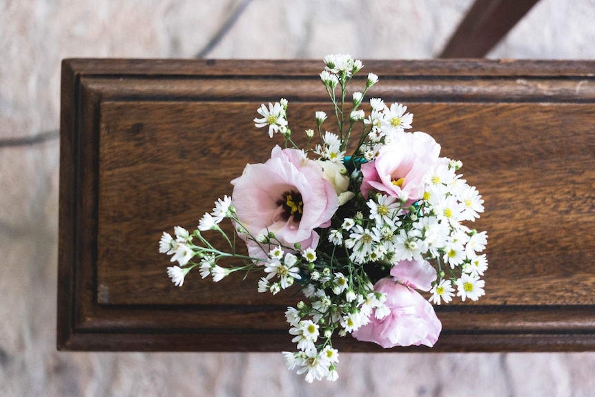 A pink and white flower arrangements rests on a small brown wooden coffin.