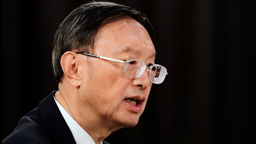 A head shot of a bespectacled man in a dark suit shows him talking.