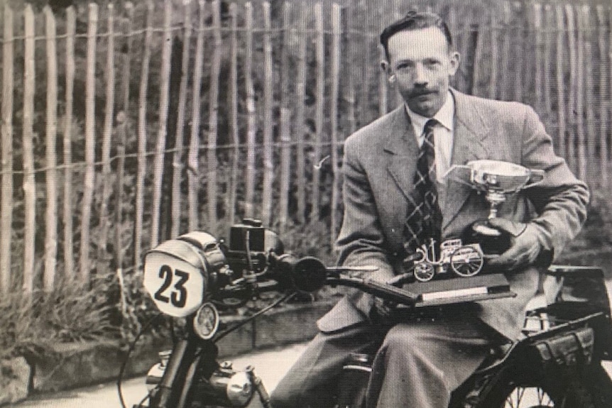 A black and white photo of a man wearing a suit and sitting on a motorbike and holding a trophy.